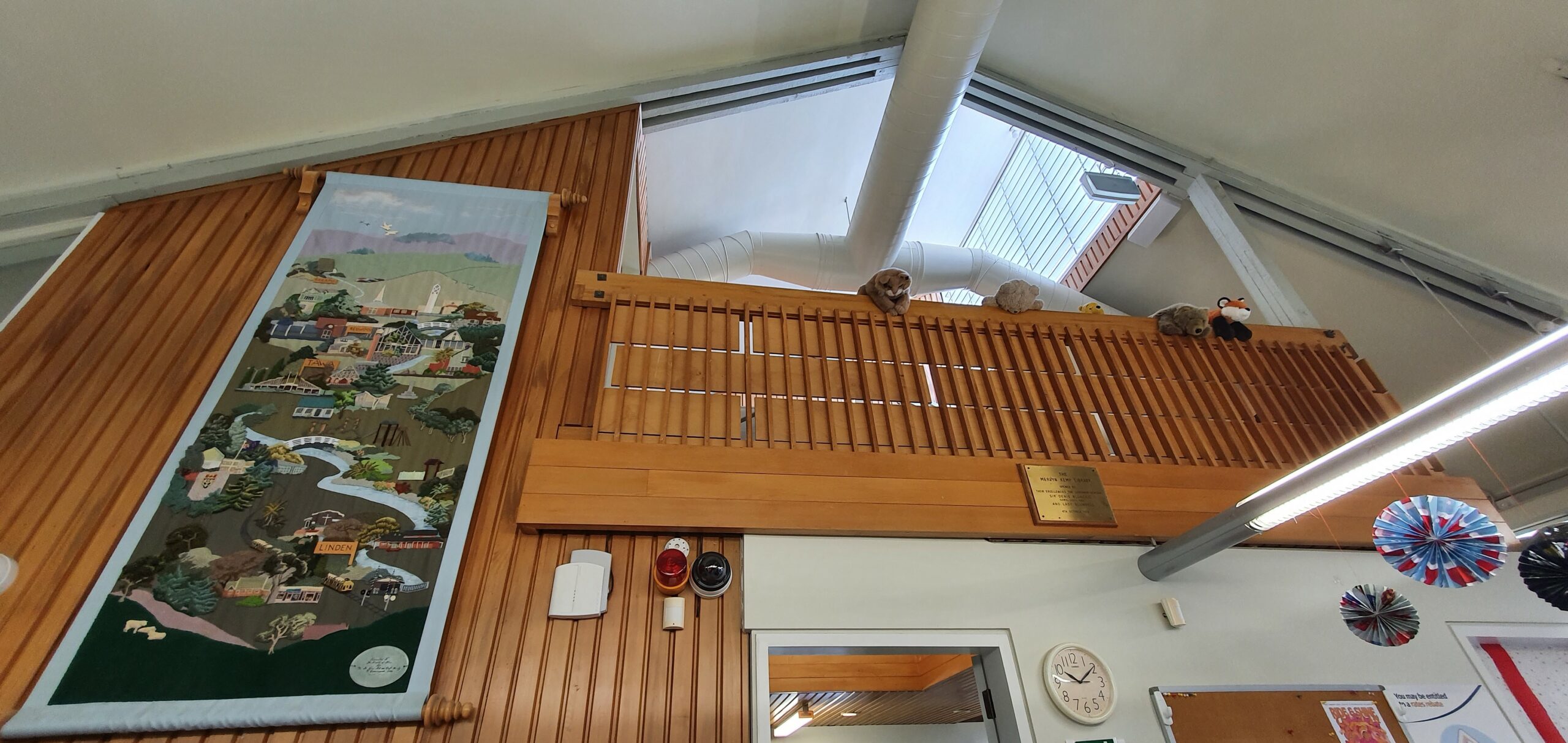 Natural wood balcony with stuffed aniimals liiking down, a skylight above, and long tapestry hanging to the left.